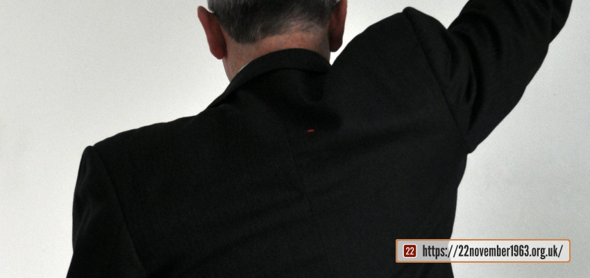 A seated figure wearing a black jacket with a red dot and raising his right arm straight up, from behind.