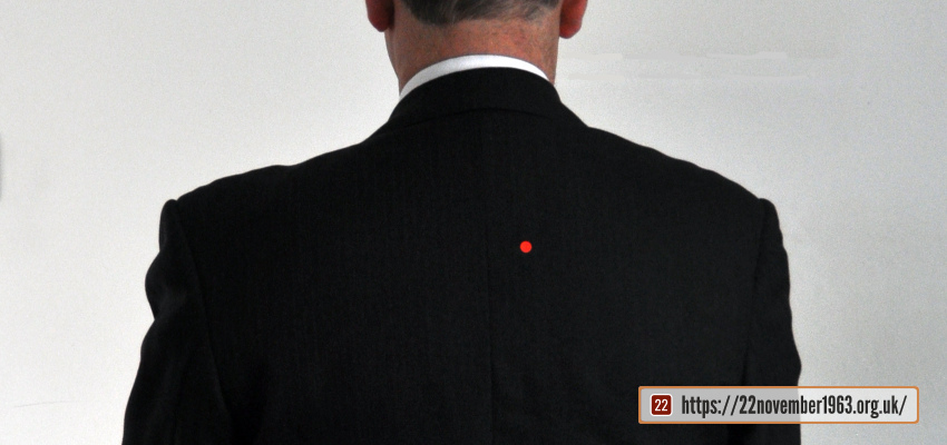 A seated figure wearing a black jacket with a red dot and raising his right arm, from behind.