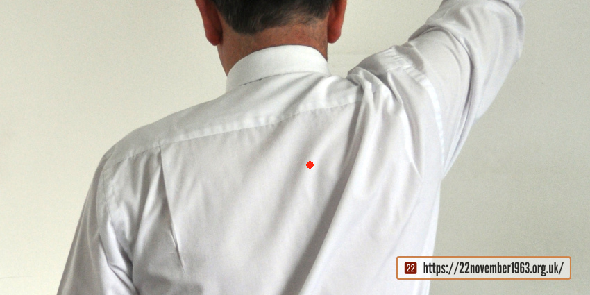 A seated figure wearing a white shirt with a red dot and raising his right arm, from behind.